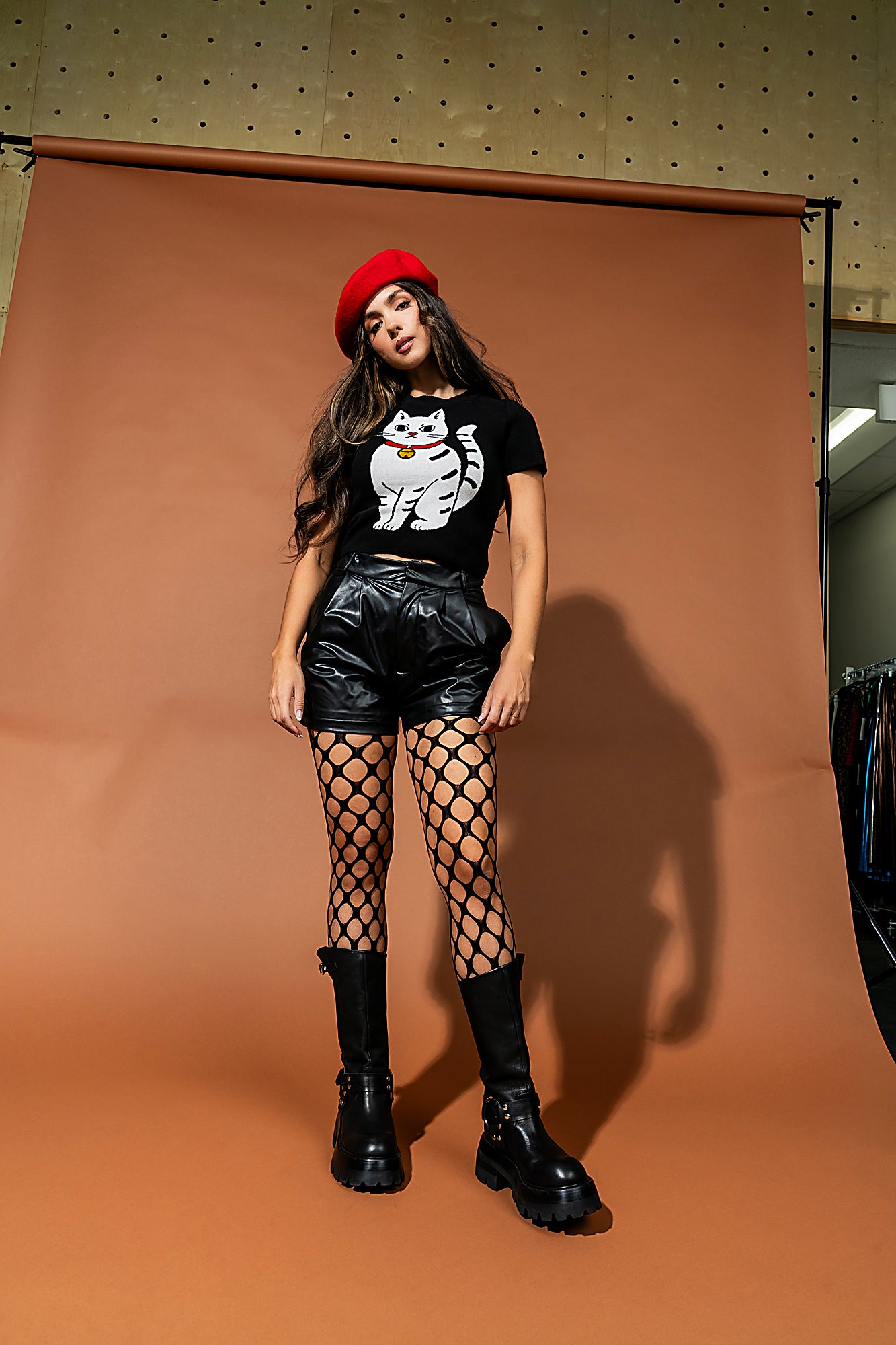 Woman posing in front of a brown backdrop wearing a black t-shirt with a white cat design, black leather shorts, fishnet stockings, and black boots.