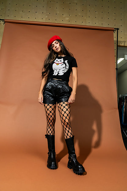 Woman posing in front of a brown backdrop wearing a black t-shirt with a white cat design, black leather shorts, fishnet stockings, and black boots.