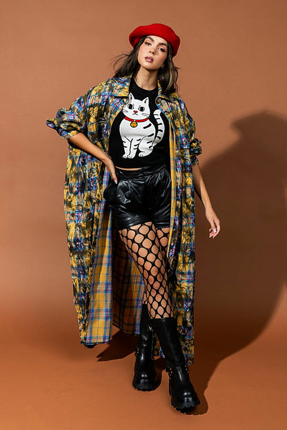 Woman in stylish outfit with plaid shawl, black top with cat graphic, leather shorts, and black boots on a brown background