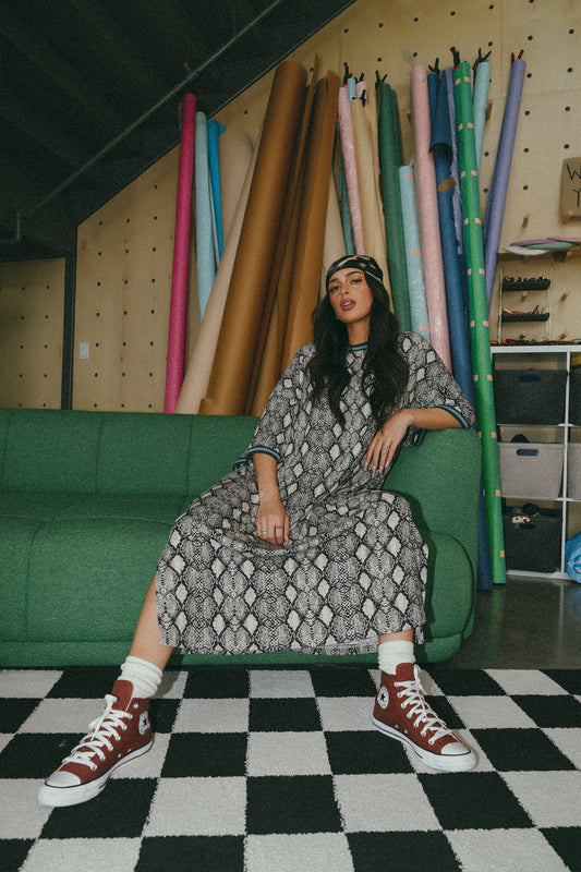 Beautiful model is seated on a green couch wearing a snake print oversized Frankie dress with a headscarf and brown high top converse shoes. 