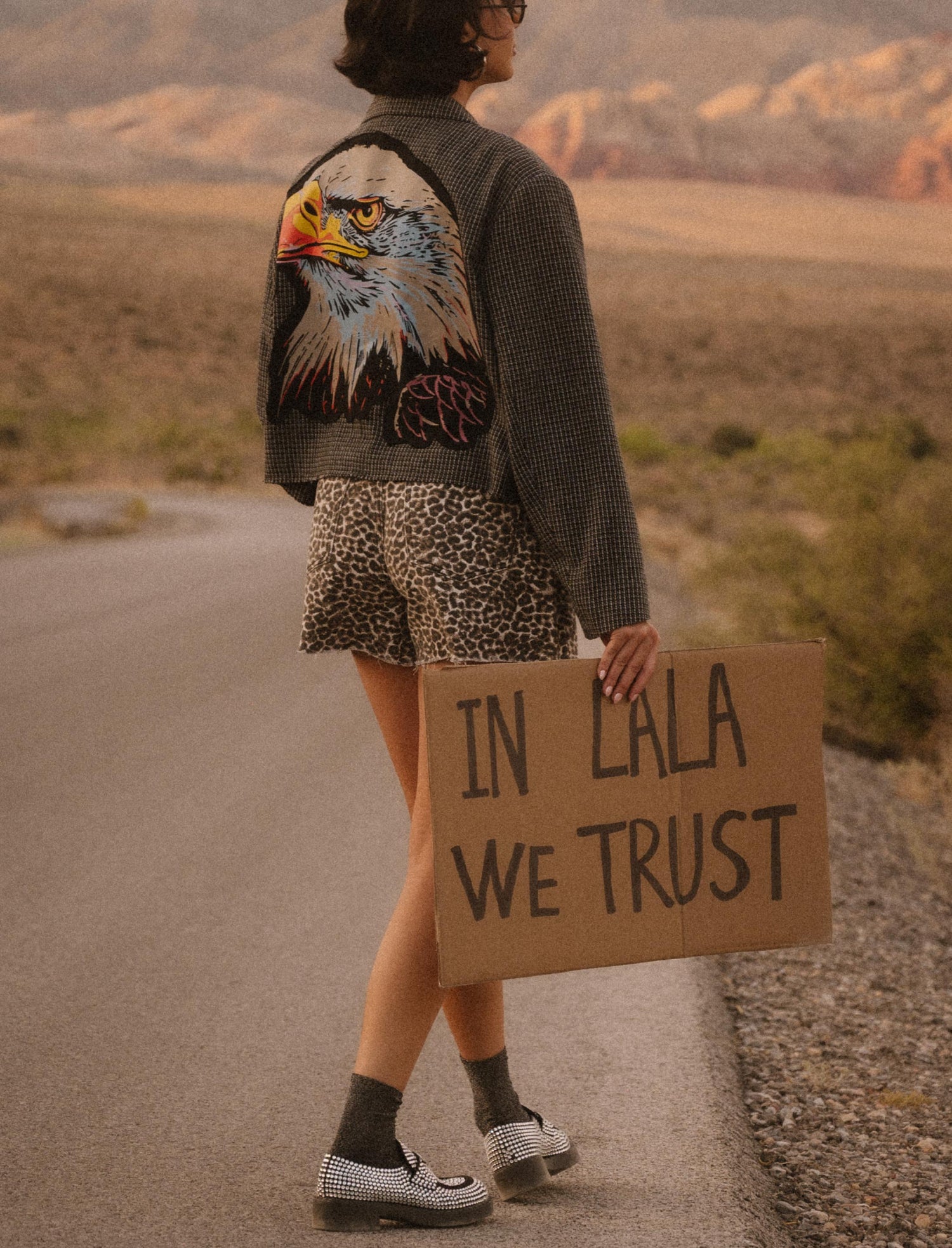Person holding a sign 'IN LALA WE TRUST' in a desert setting