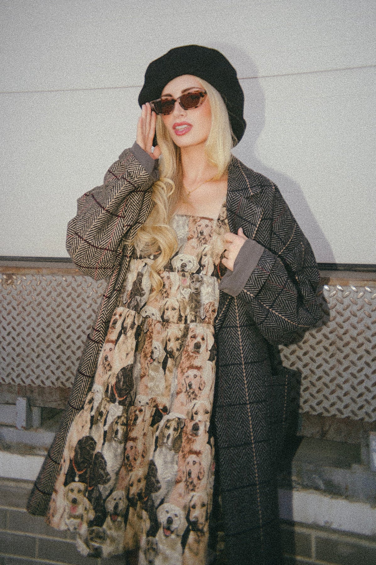 Woman wearing a patterned coat and scarf with a black beret and sunglasses, standing against a light-colored wall.