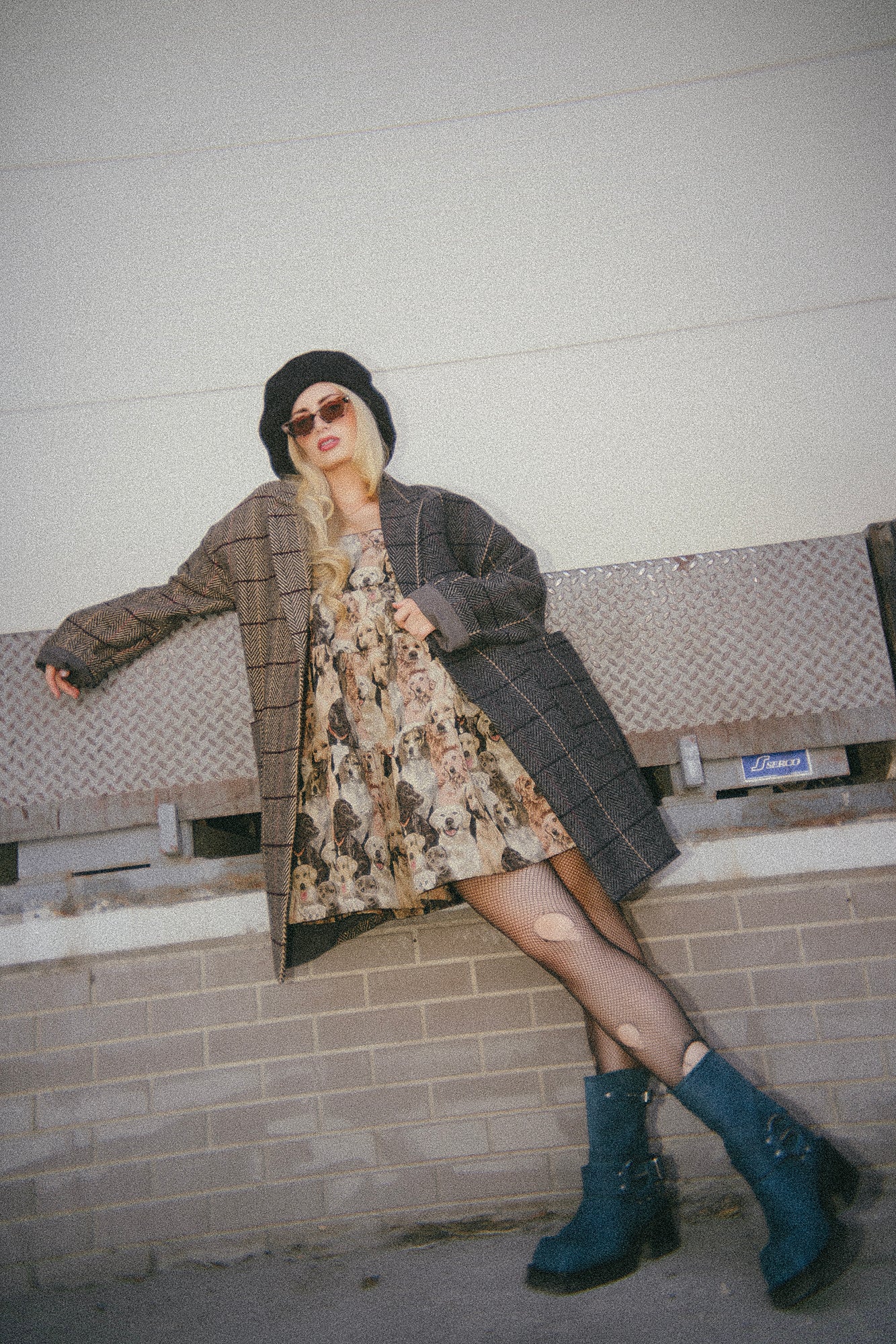 Person wearing a long coat, dog dress, and blue boots against a plain wall.