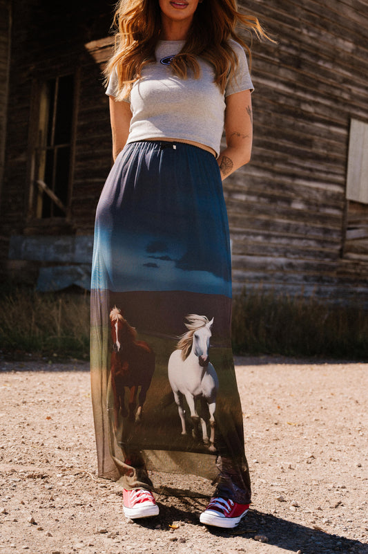 Woman wearing a long skirt with horse design in front of a wooden building.