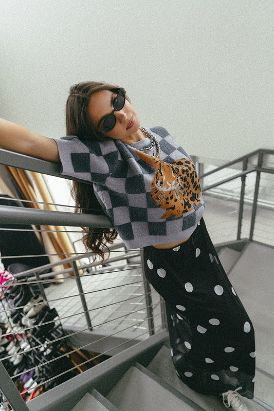 Woman leaning on a railing wearing a grey checkered knit top with a leopard print bunny and black mesh maxi skirt with white polka dots.