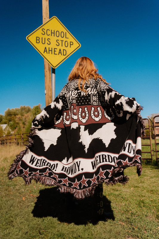 Person wearing a large black and white patterned cardigan with text, standing in front of a 'School Bus Stop Ahead' sign.