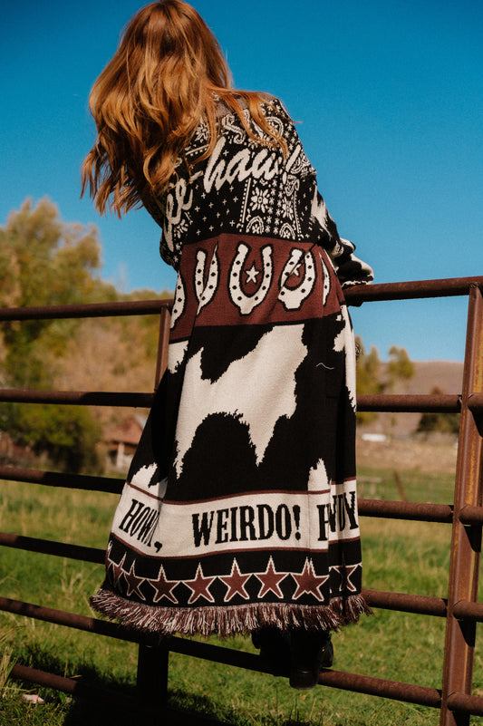 Person wearing a patterned cardigan with text and graphics, standing by a fence in an outdoor setting.
