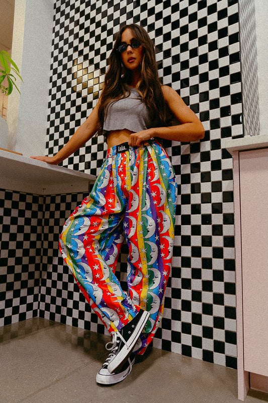 Person wearing pants with rainbow stripes, moons and stars standing in front of a checkered wall.