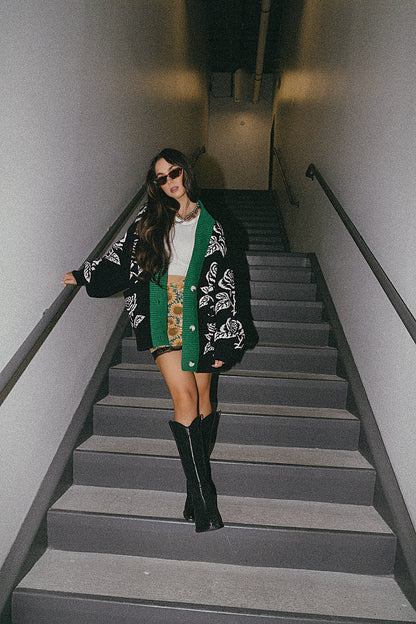 Woman walking down a set of stairs wearing a black and white floral cardigan, green scarf, and black knee-high boots.