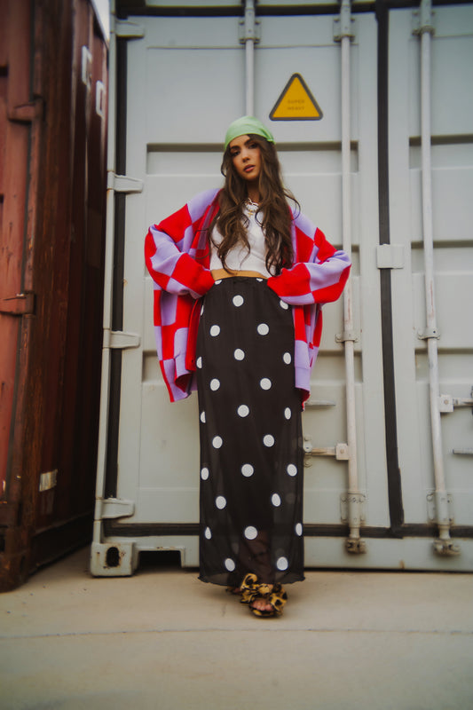 Woman in red and lavender checkerboard cardigan and black mesh maxi skirt with white polka dots standing in front of shipping containers