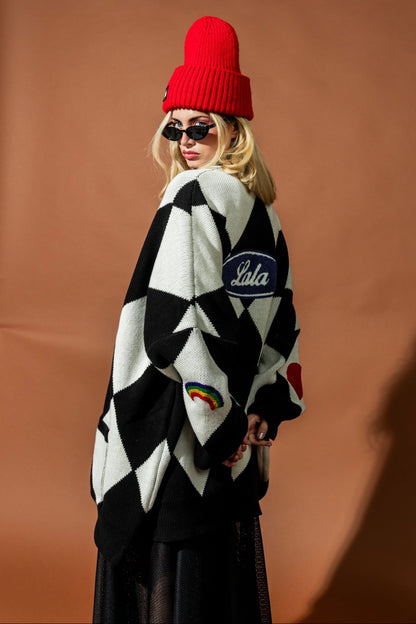 Person wearing a black and white patterned cardigan with 'Lala' branding, red beanie, and sunglasses on a brown background