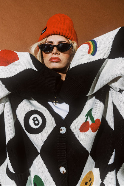 Person wearing a black and white patterned cardigan with colorful designs, sunglasses, and an orange beanie.