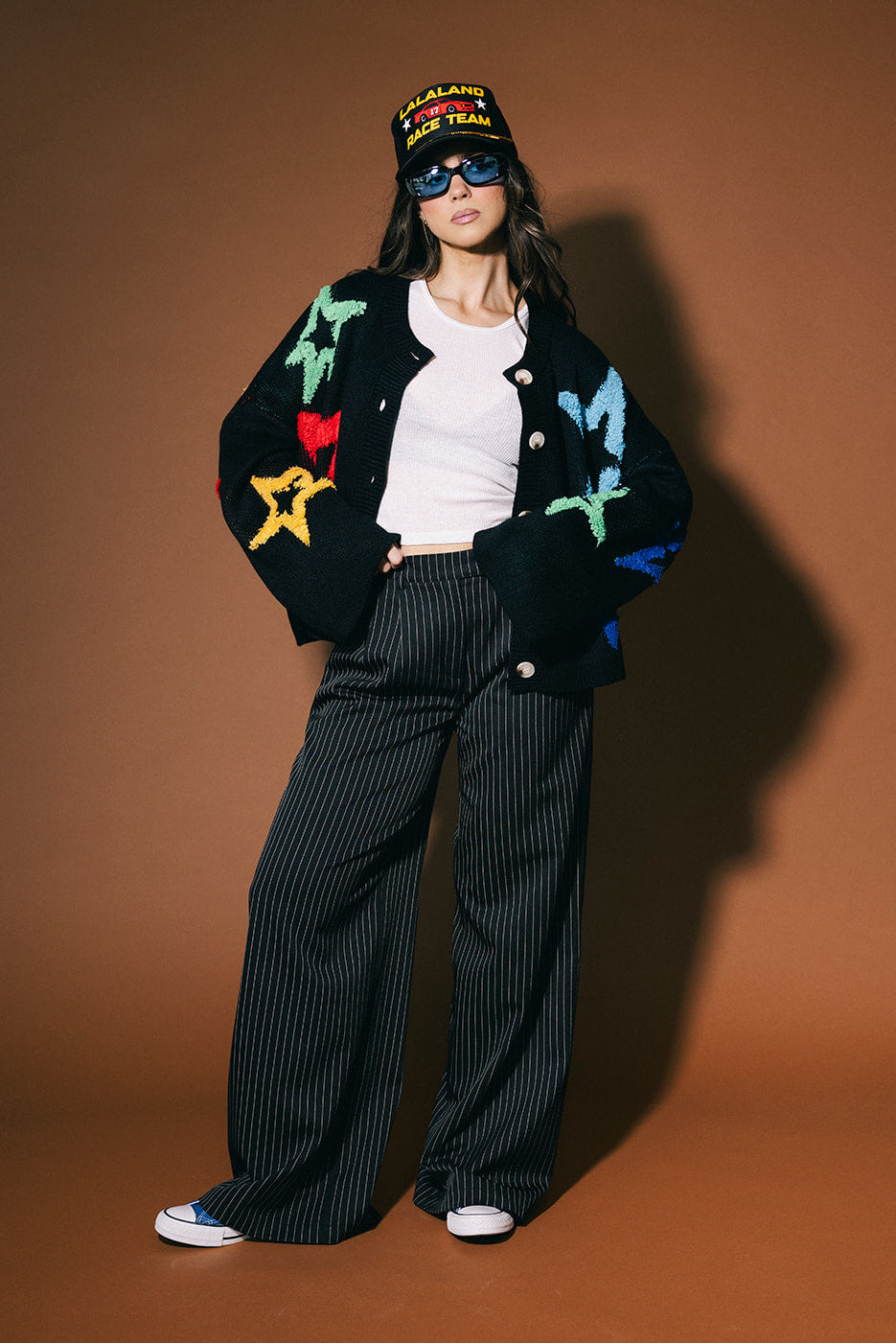 Person wearing a colorful jacket with star designs, black pants, and a cap on a brown background