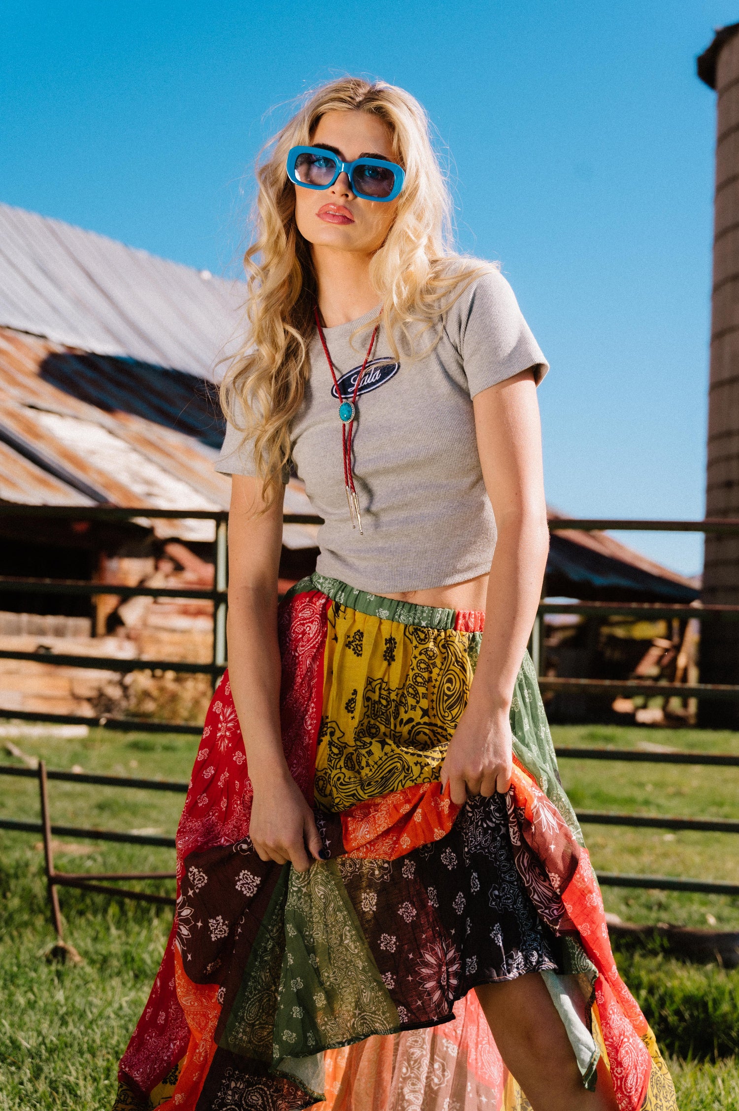 Woman wearing a grey ribbed baby tee with a 'Lala' logo, sunglasses, and a colorful skirt standing in front of a wooden barn.