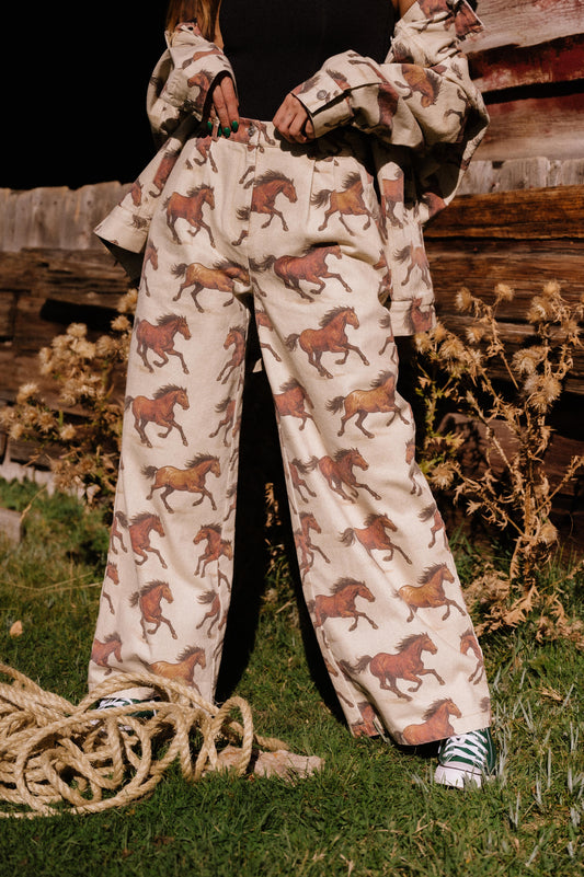 Person wearing white denim wide leg trousers with a horse pattern outdoors with a wooden fence and plants in the background