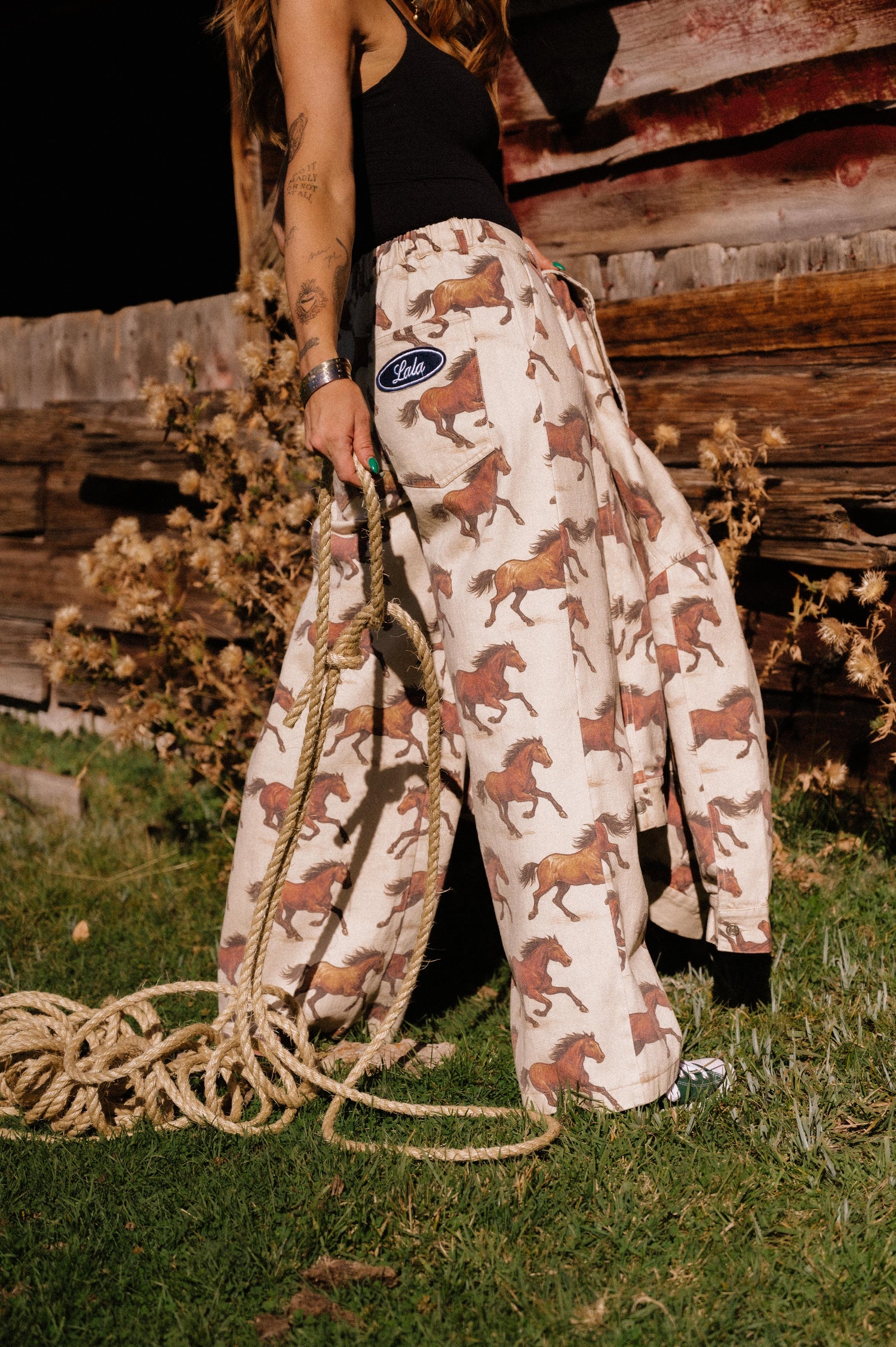 Person wearing white denim wide leg trousers with a horse pattern holding a rope in front of a wooden barn.