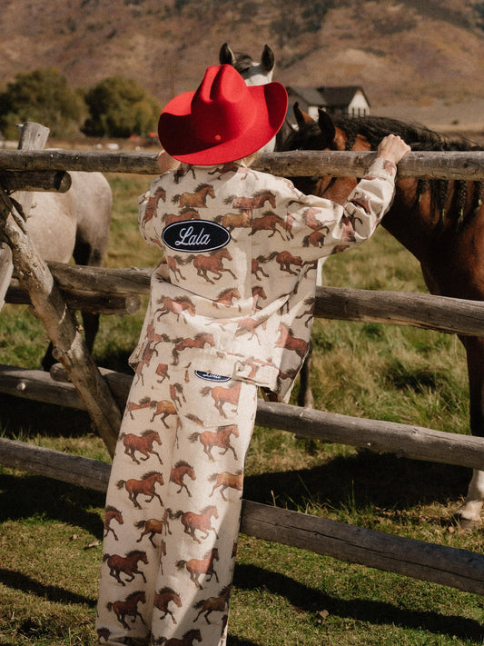 Person wearing a red cowboy hat and white denim jacket with a horse pattern standing next to a wooden fence with horses in the background.