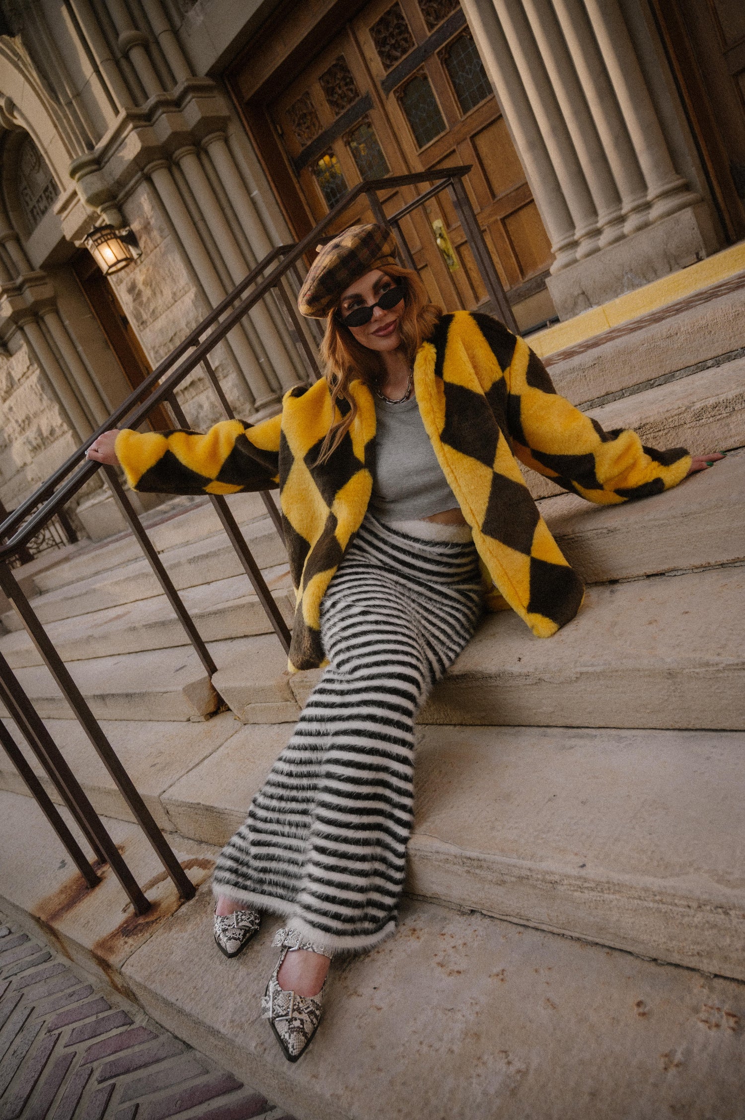 Woman in a yellow and brown diamond faux fur coat and black and white striped maxi skirt with fuzzy texture sitting on steps in front of a building.