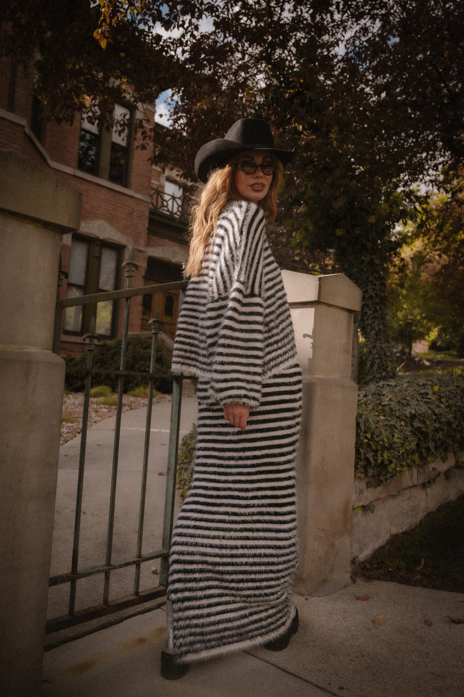 Person wearing a black and white striped sweater and maxi skirt with fuzzy texture with a cowboy hat in front of a fence.