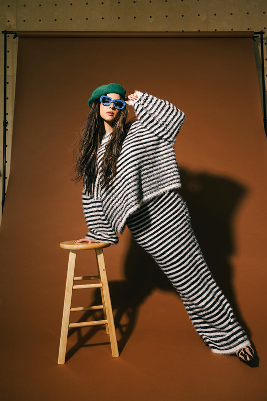 Person wearing a black and white striped sweater and skirt with a green beret and blue sunglasses, standing next to a wooden stool against a brown background.