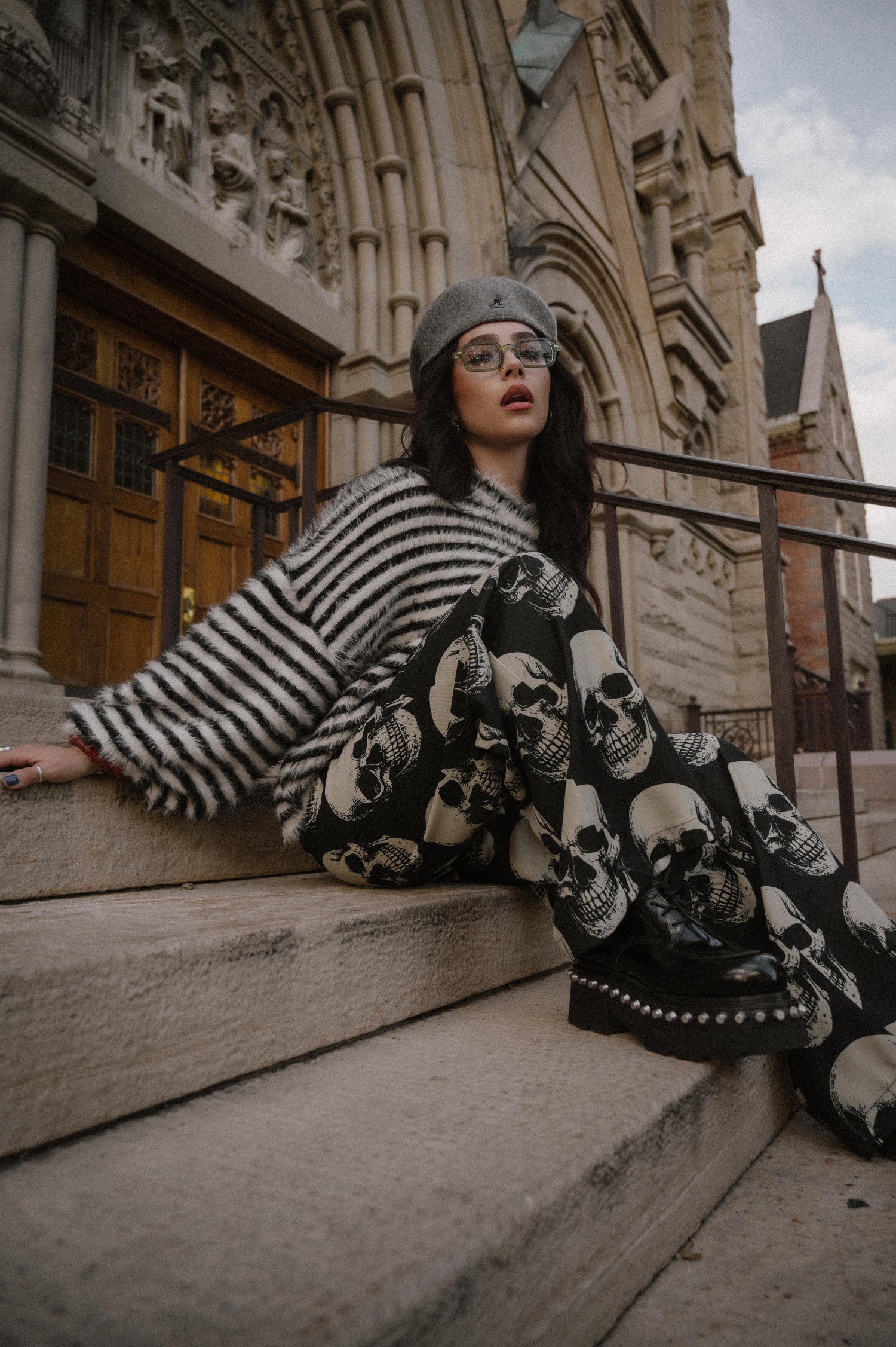 Woman sitting on steps wearing a black and white striped sweater with skull pattern trousers, in front of a large building.