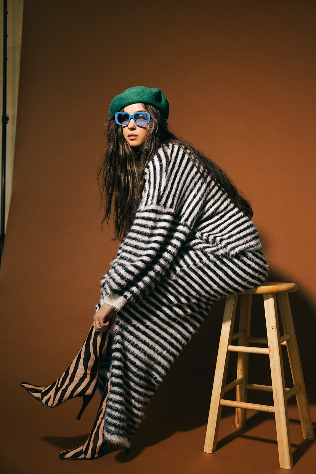 Woman in a striped sweater and skirt sitting on a stool against a brown background