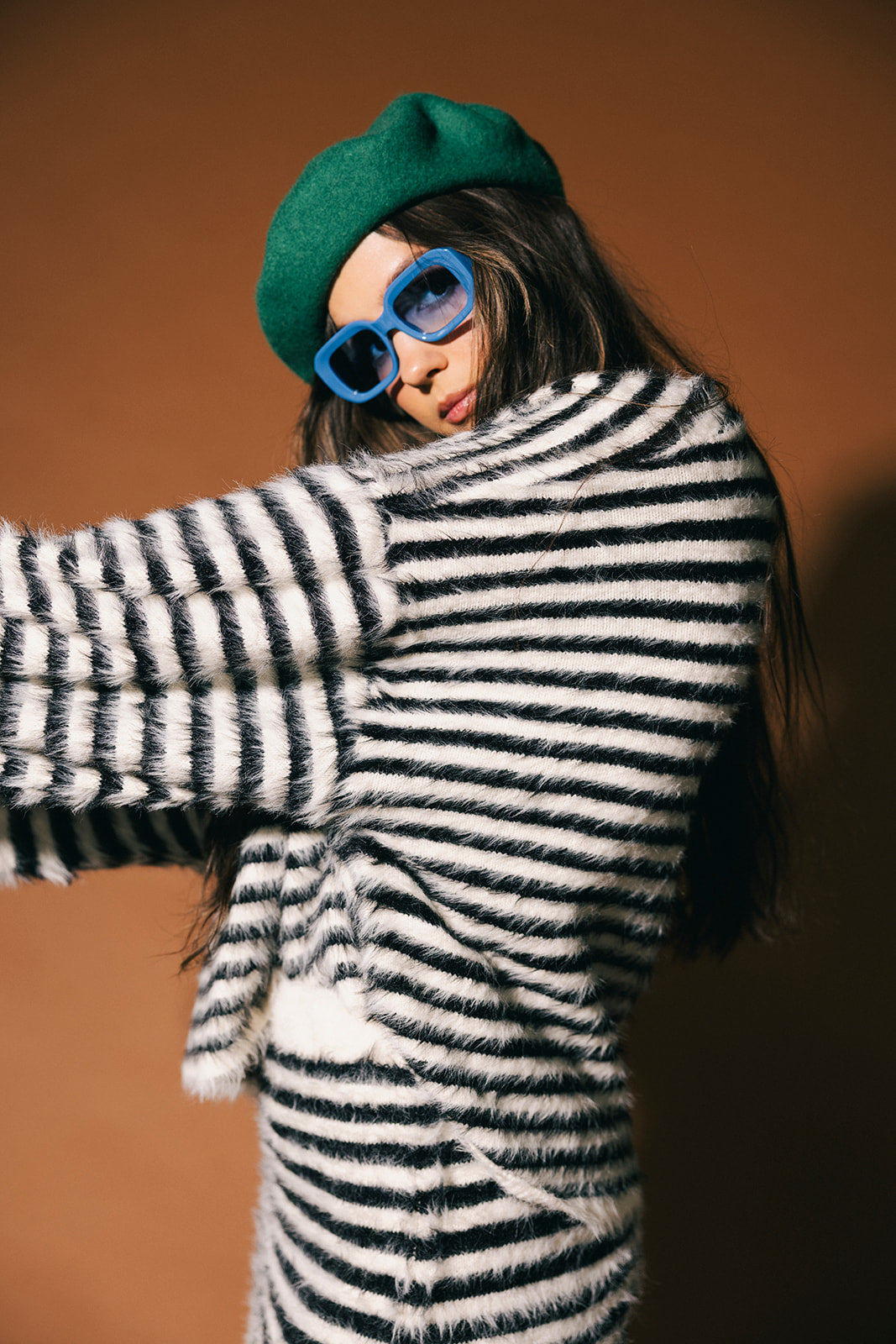 Person wearing a black and white striped fuzzy texture sweater with a green beret and blue sunglasses against a brown background