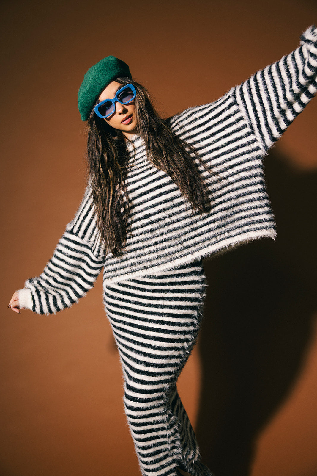 Person wearing a black and white striped sweater and skirt with fuzzy texture with a green beret and blue sunglasses on a brown background