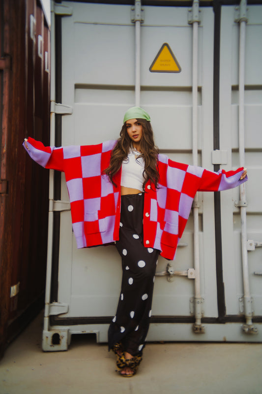 Person wearing a red and lavender checkerboard cardigan and black mesh maxi skirt with white polka dots in front of a shipping container.