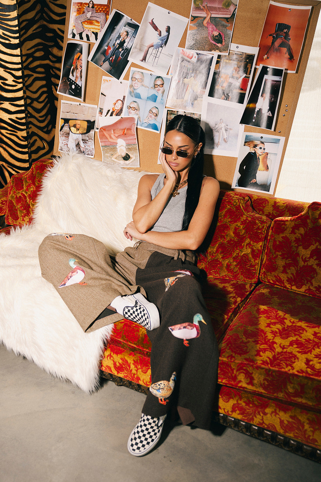 Woman sitting on a colorful couch with a collage of photos on the wall behind her wearing half and half trousers with duck patches.