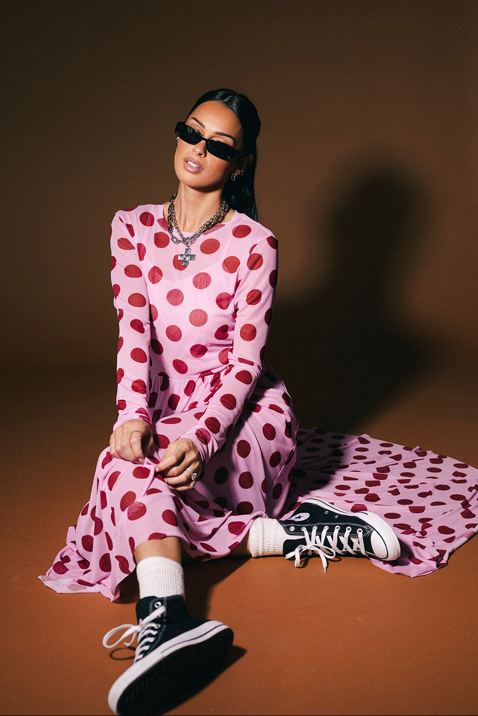 Woman wearing a pink polka dot dress and black sneakers sitting on a brown background