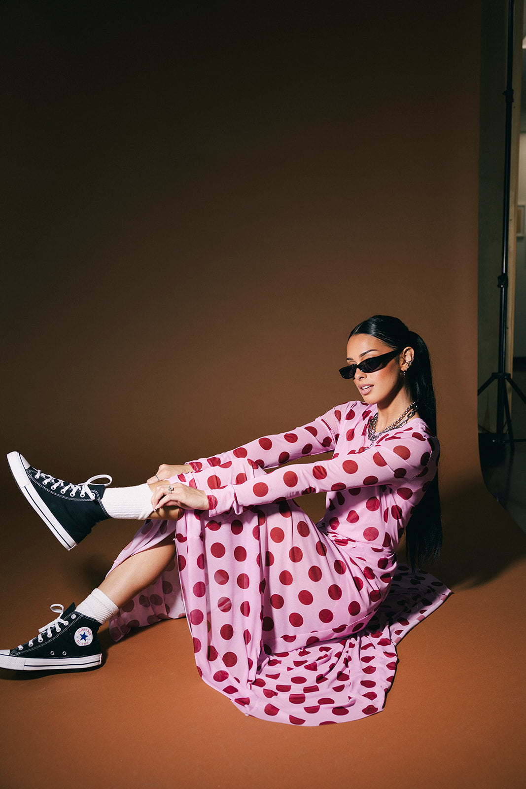 Woman in a pink polka dot dress and black sneakers sitting on a brown floor.