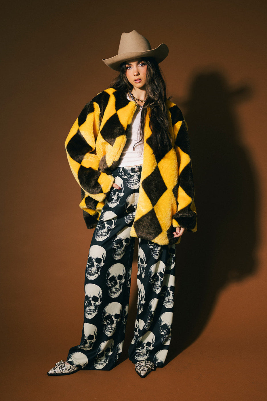 Person wearing a yellow and brown diamond faux fur coat with skull-patterned trousers on a brown background
