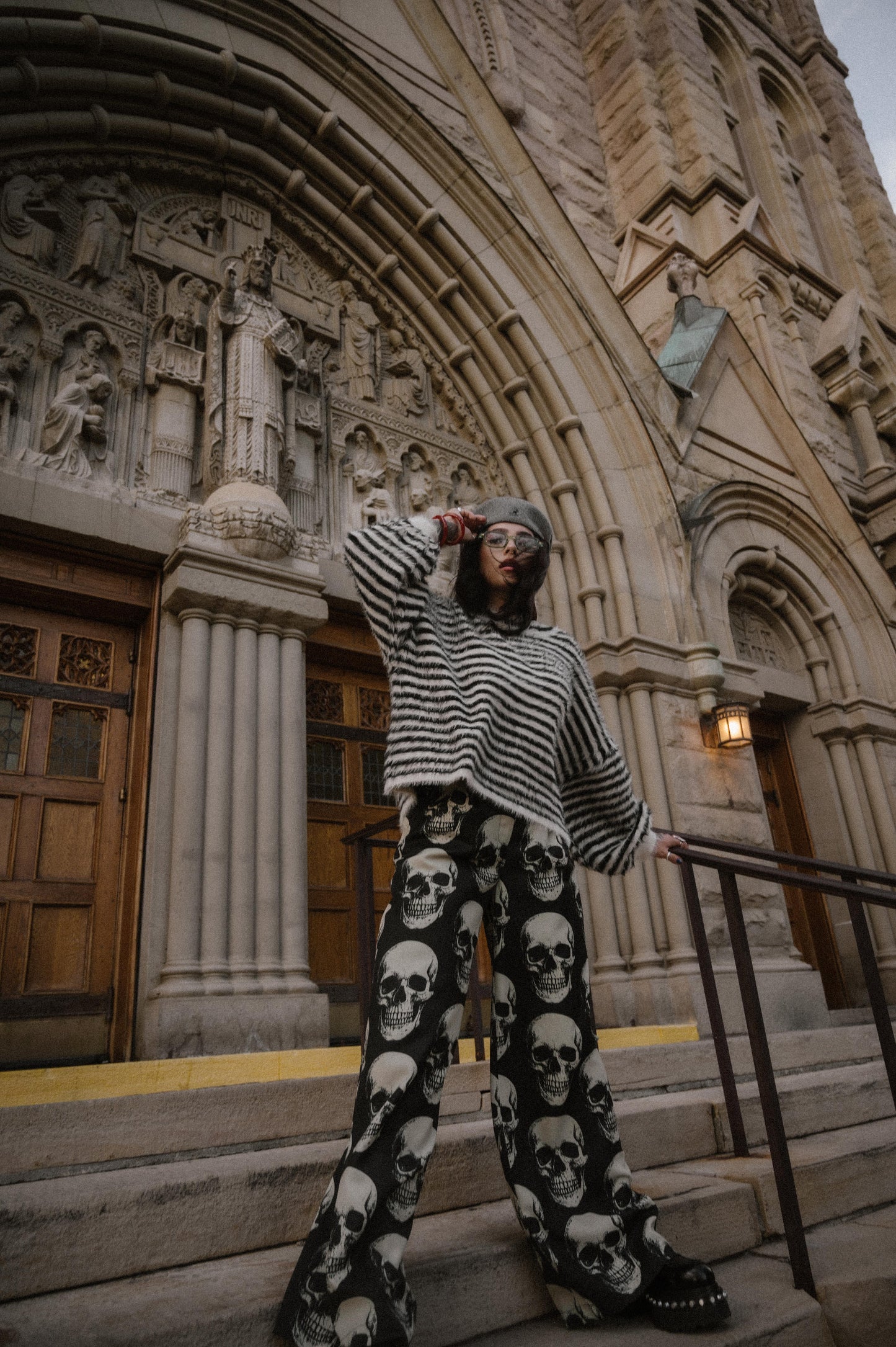 Person wearing a black and white striped sweater and skull-patterned trousers standing on steps in front of a large building with architectural details.