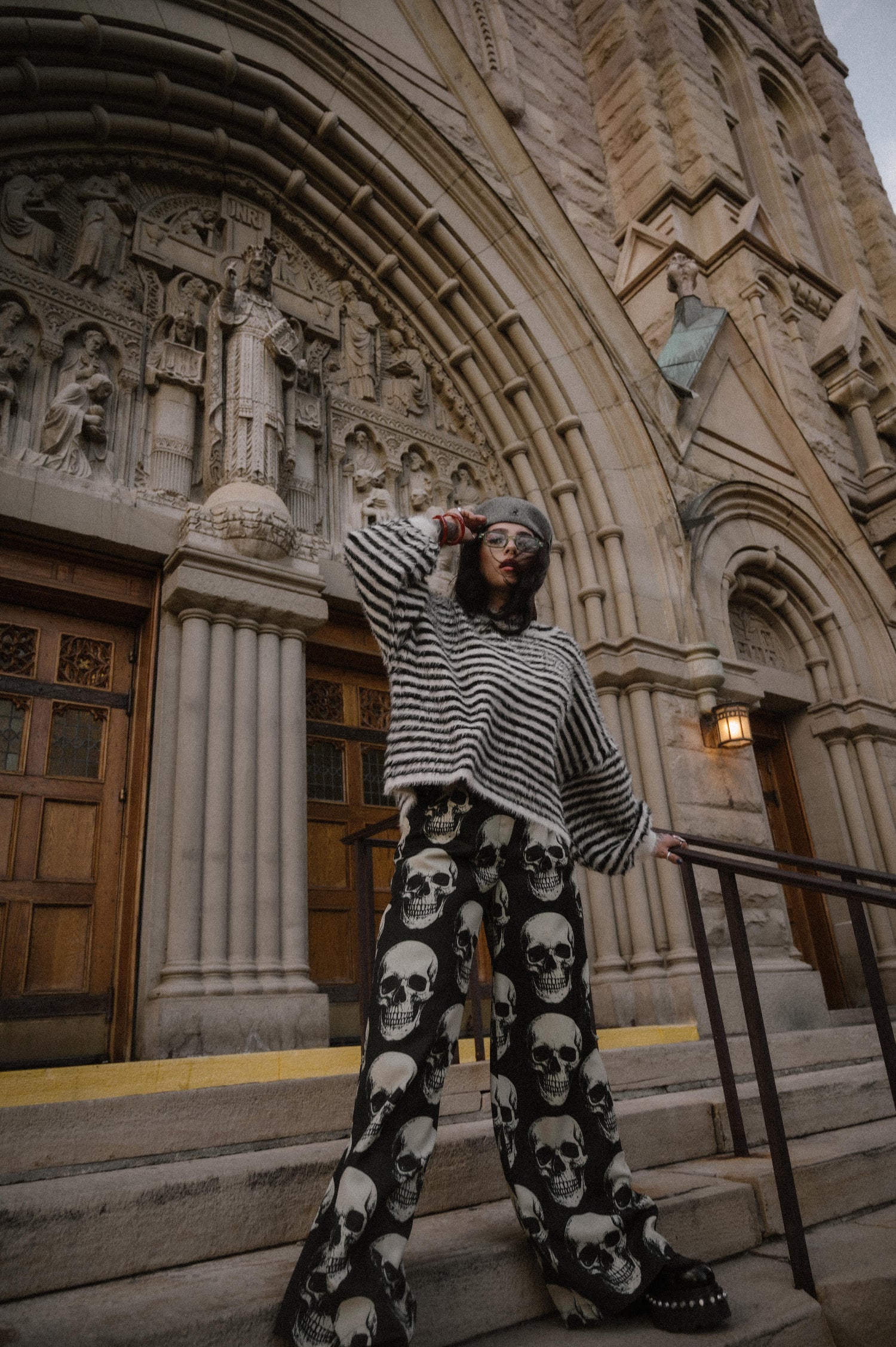 Person wearing a black and white striped sweater and skull-patterned trousers standing on steps in front of a large building with architectural details.