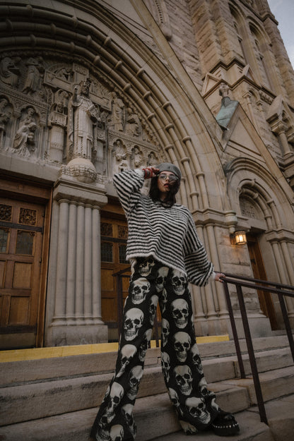 Person wearing a black and white striped sweater and skull-patterned trousers standing on steps in front of a large building with architectural details.