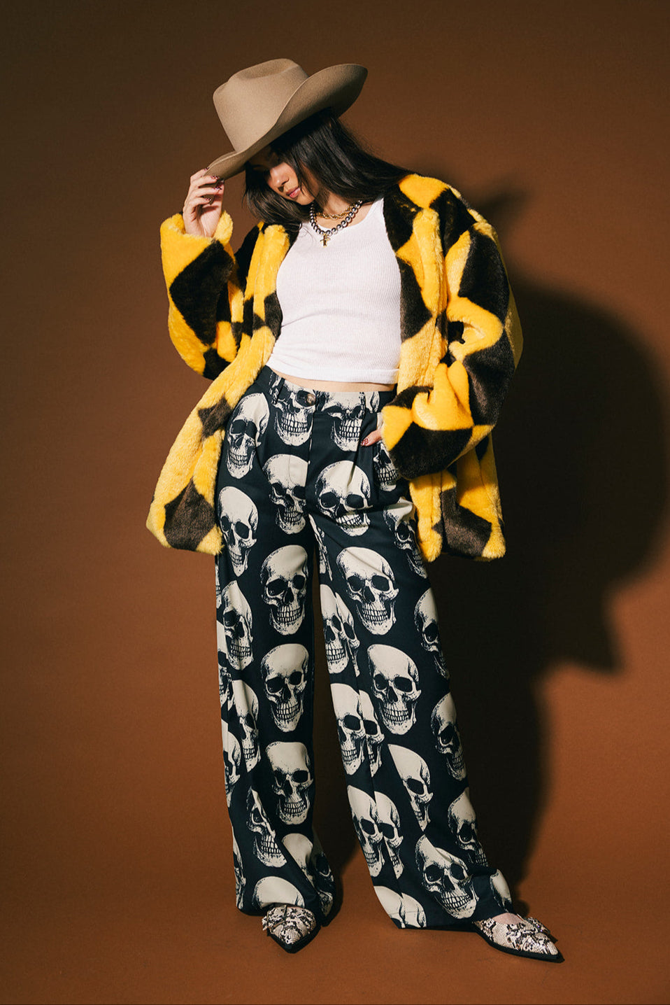 Person wearing a yellow and brown diamond faux fur coat, white top, and skull-patterned trousers against a brown background.