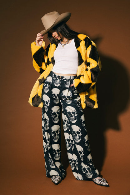 Person wearing a yellow and brown diamond faux fur coat, white top, and skull-patterned trousers against a brown background.