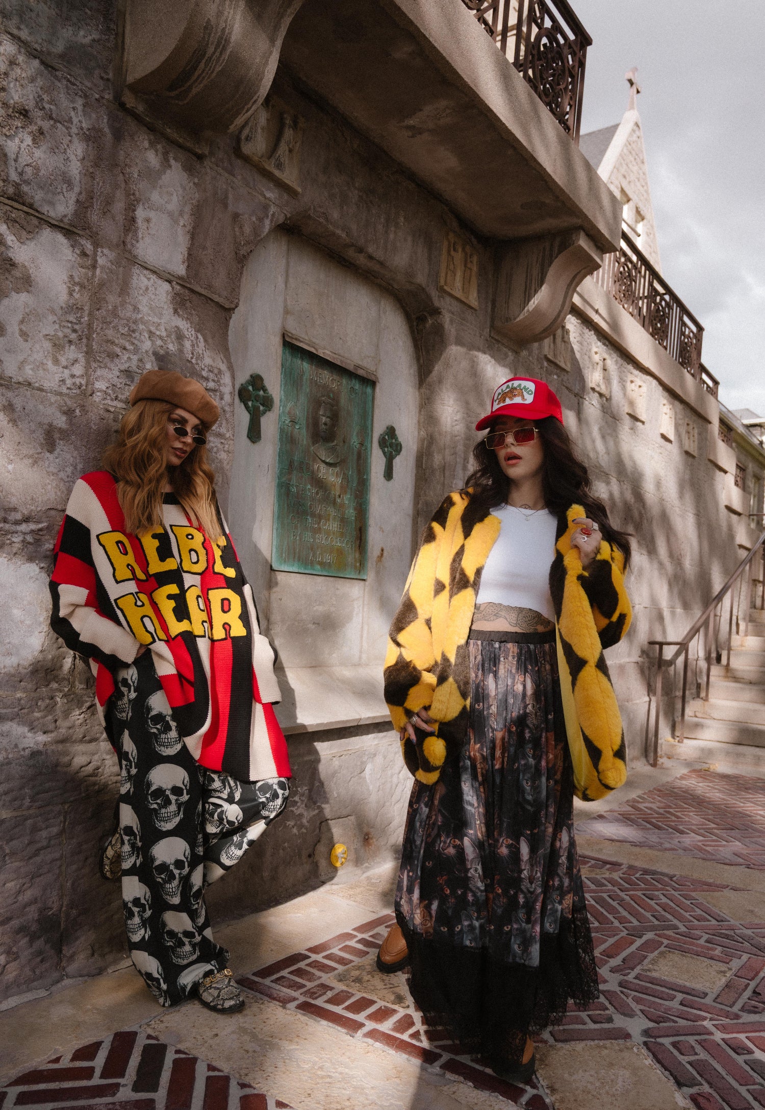 Two women standing on a street with a building in the background. One is wearing black trousers with skull pattern.