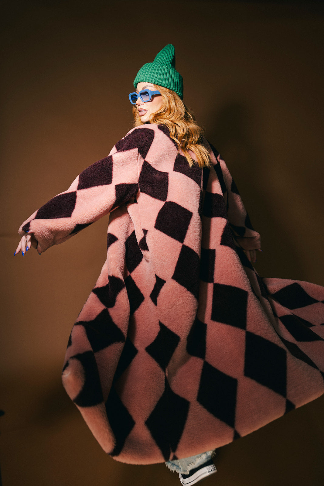 Person wearing a pink and maroon harlequin long faux fur coat on a brown background