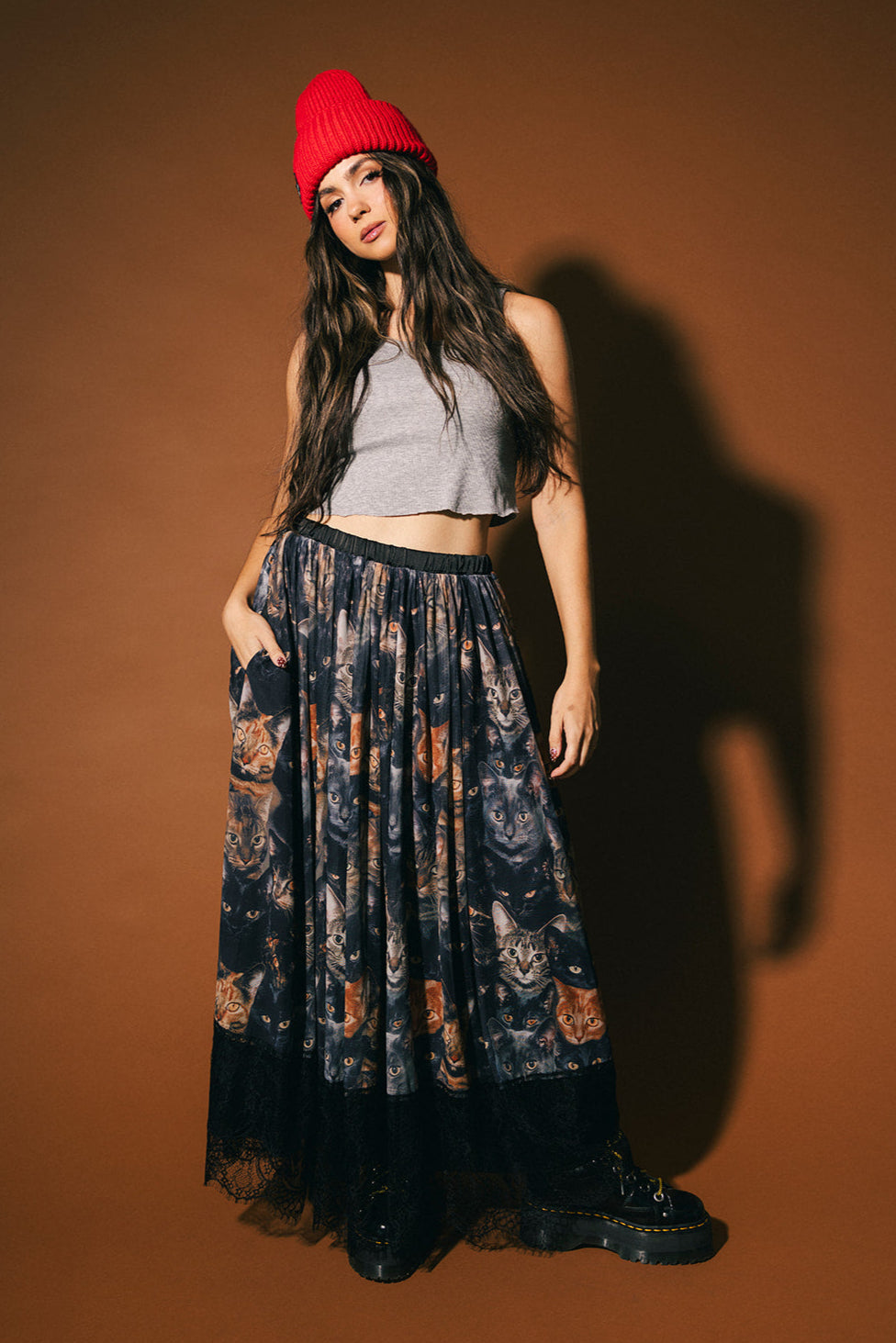 Woman wearing a red beanie, gray crop top, and cat patterned mesh skirt with lace hem against a brown background