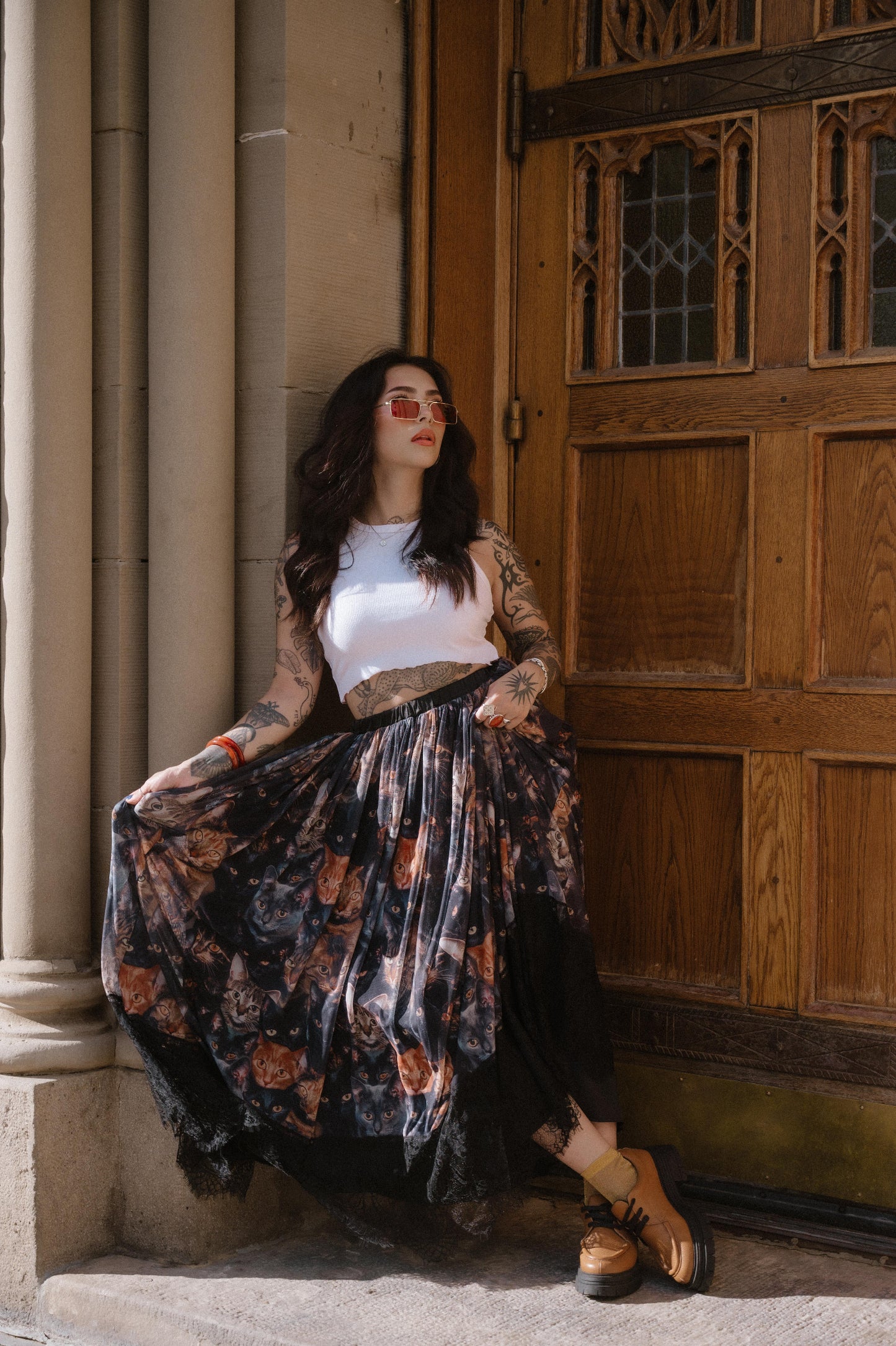 Woman leaning against a stone wall with a wooden door in the background wearing a cat pattern mesh maxi skirt with lace hem.