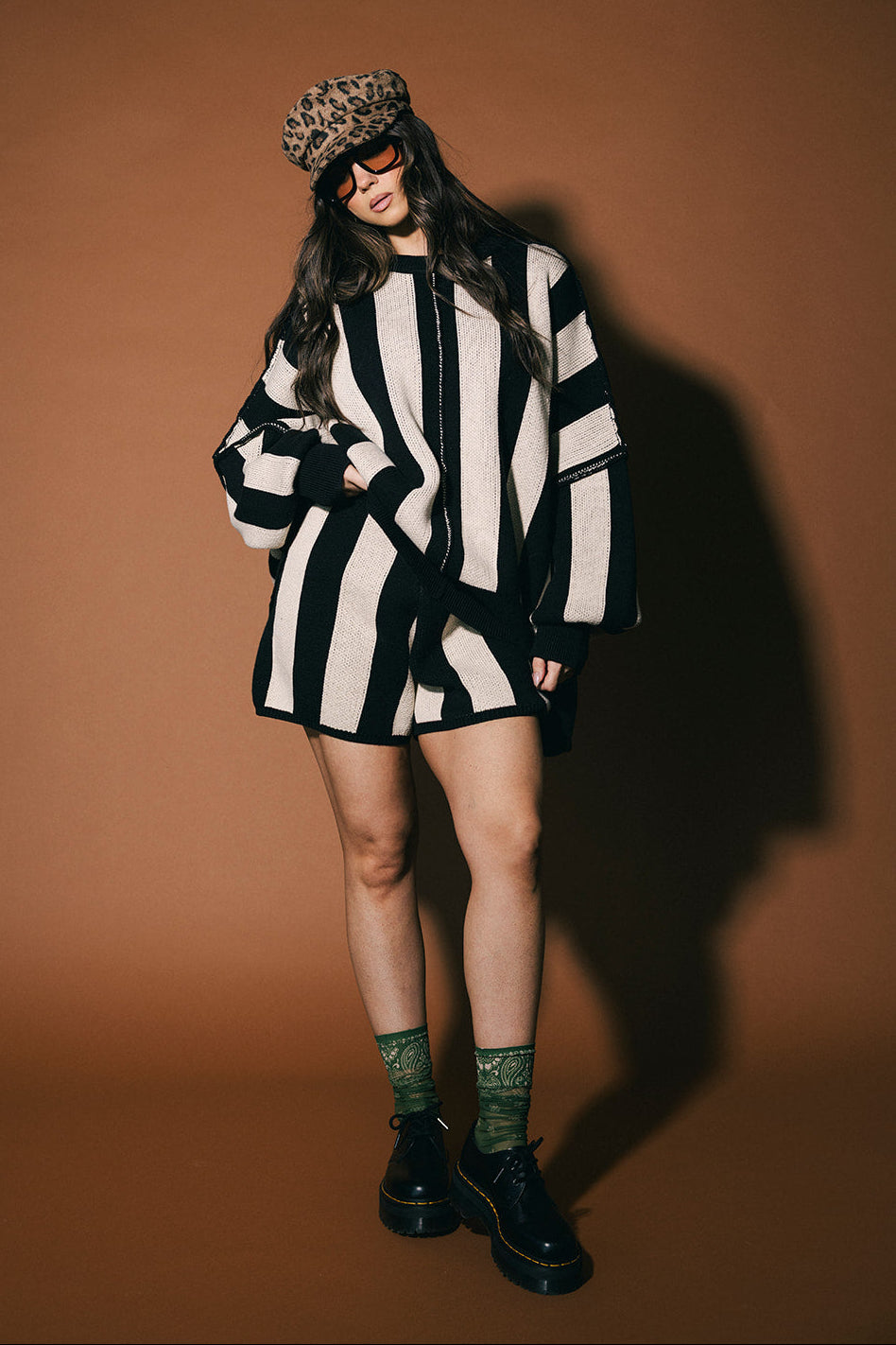 Person wearing a black and white striped outfit with a leopard print hat against a brown background