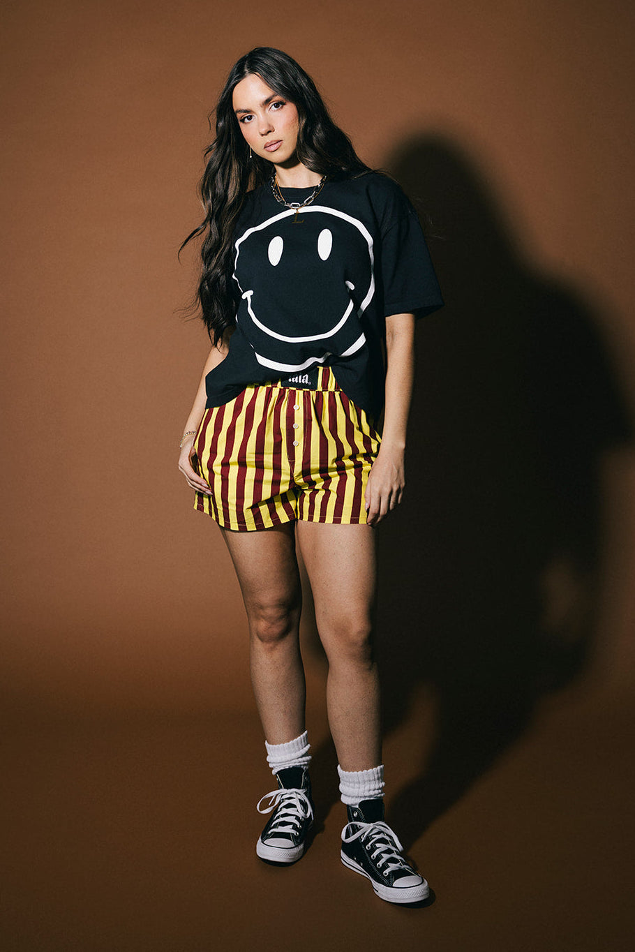 Person wearing a black t-shirt with a smiley face design, yellow and red striped shorts, and black sneakers on a brown background.
