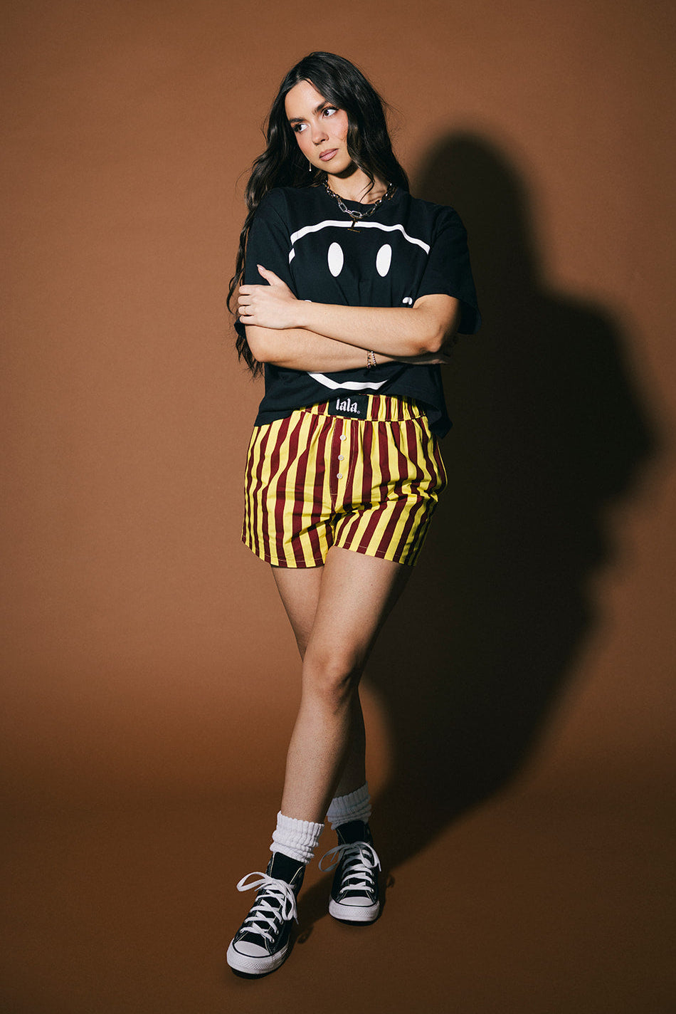 Person wearing a black t-shirt with a smiley face design, yellow and red striped shorts, and black sneakers on a brown background.