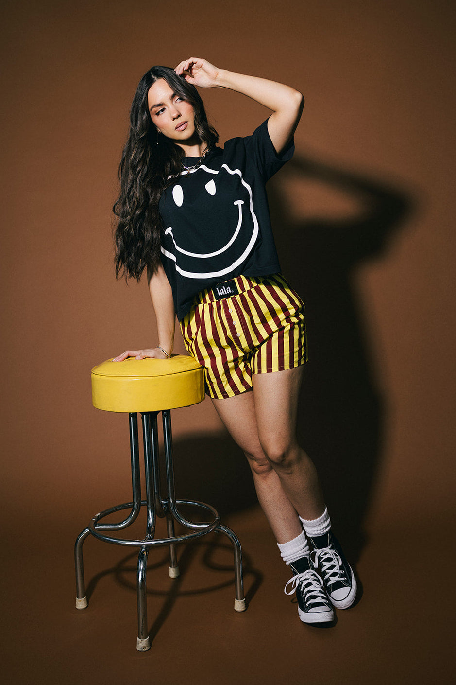 Person wearing a black smiley face shirt and yellow and red striped shorts standing next to a yellow stool on a brown background.