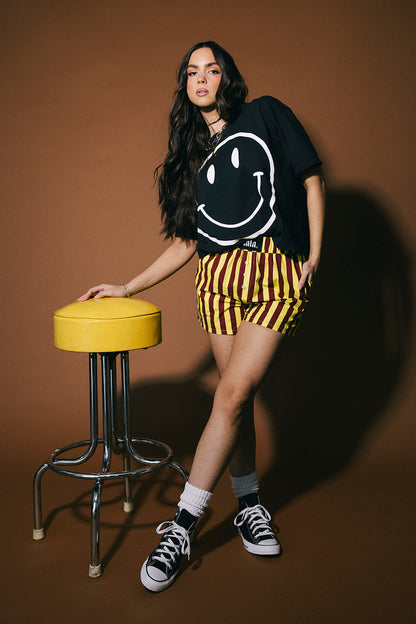 Woman in black smiley face t-shirt and yellow striped shorts standing next to a yellow stool on a brown background