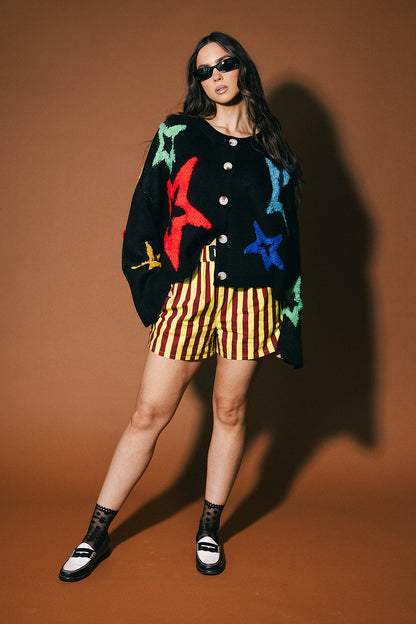 Woman wearing a colorful cardigan, striped shorts, and sunglasses on a brown background