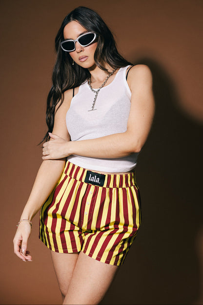 Woman wearing a white sleeveless top and red and yellow striped shorts with 'lala' branding on a brown background.