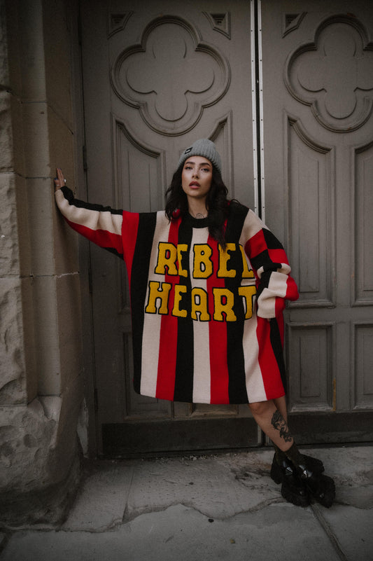 Person wearing an oversized striped sweater with 'Rebel Heart' text standing against a stone wall.